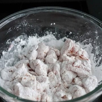 Cubed chicken thighs being tossed and coated evenly in white cornstarch powder inside a mixing bowl.