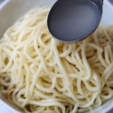 Cooked noodles placed over sauce in a large bowl with noodle cooking water being added.