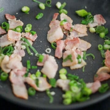Bacon cooking in a pan with garlic and green onions being sautéed until fragrant.