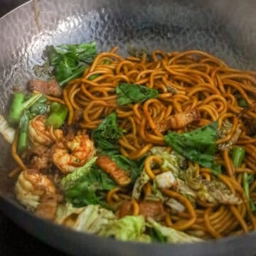 Stir-fried Hokkien noodles with shrimp, pork, cabbage, and broccoli greens in a wok.