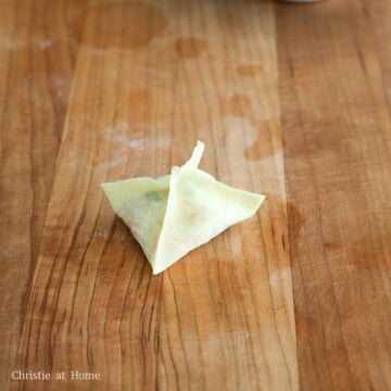Hands pressing the remaining side seams of a folded wonton wrapper together to completely close the pouch.