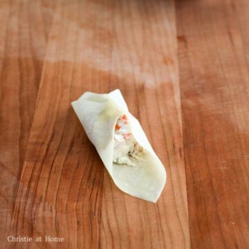 Two fingers pinching two opposite corners of a square wonton wrapper together over the cream cheese filling.