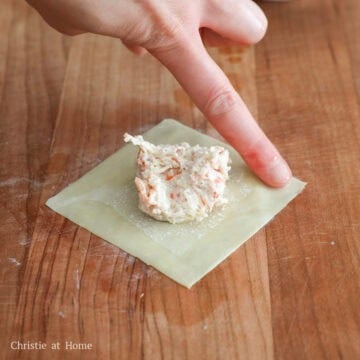 A teaspoon placing crab filling onto the center of a flat square wonton wrapper next to a small bowl of water.