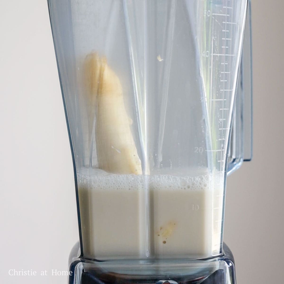 A blender filled with a peeled banana, sugar, vanilla extract, and oat milk.