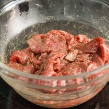 Thinly sliced flank steak being tossed with savory marinade ingredients inside a large glass mixing bowl.