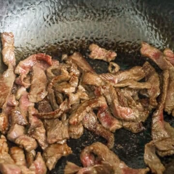 Marinated strips of flank steak searing in a single layer inside a hot, oiled skillet.