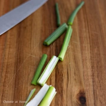 Close up shot of fresh green onions being sliced into two inch pieces on a wooden cutting board.