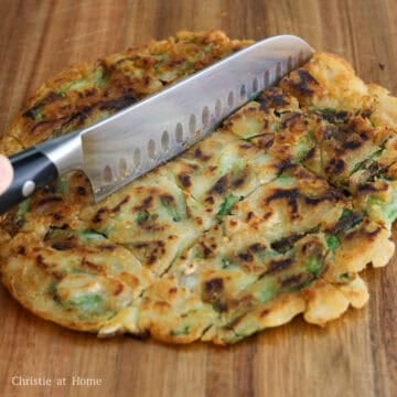 A sharp knife slicing a crispy green onion pancake into bite sized squares on a cutting board.