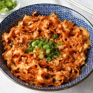 Noodles being tossed with chopsticks until evenly coated in sauce, finished with green onions and sesame seeds.