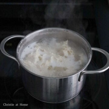 Boiling wheat noodles in a pot of water, then draining and transferring them to a large heatproof bowl.