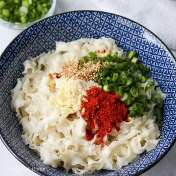 Cooked noodles in a bowl with soy sauce, dark soy sauce, black vinegar, garlic, bouillon powder, sugar, green onions, chili flakes, and sesame seeds placed in the center.