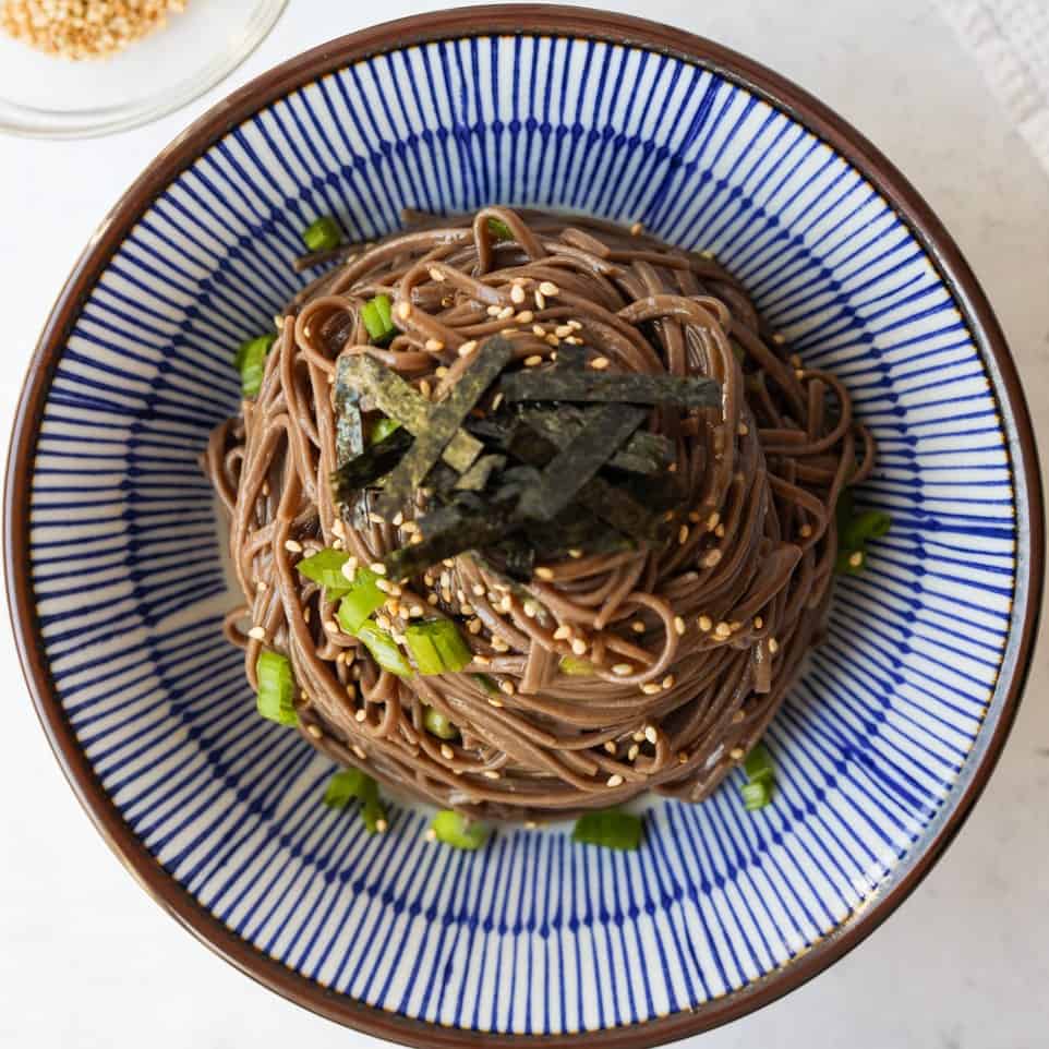 A serving of cold sesame soba noodles in a bowl, garnished with thin strips of roasted seaweed on top.