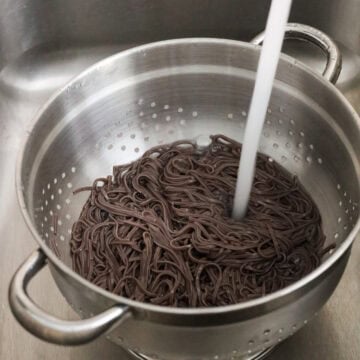 Cooked soba noodles being rinsed under cold running tap water inside a colander.