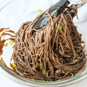 A bowl filled with buckwheat noodles being tossed with sesame seeds, green onions, and a dark dressing.