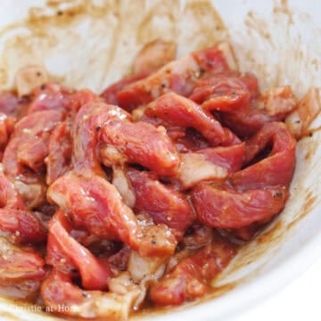 Sliced flank steak being tossed with savory marinade ingredients in a large mixing bowl.